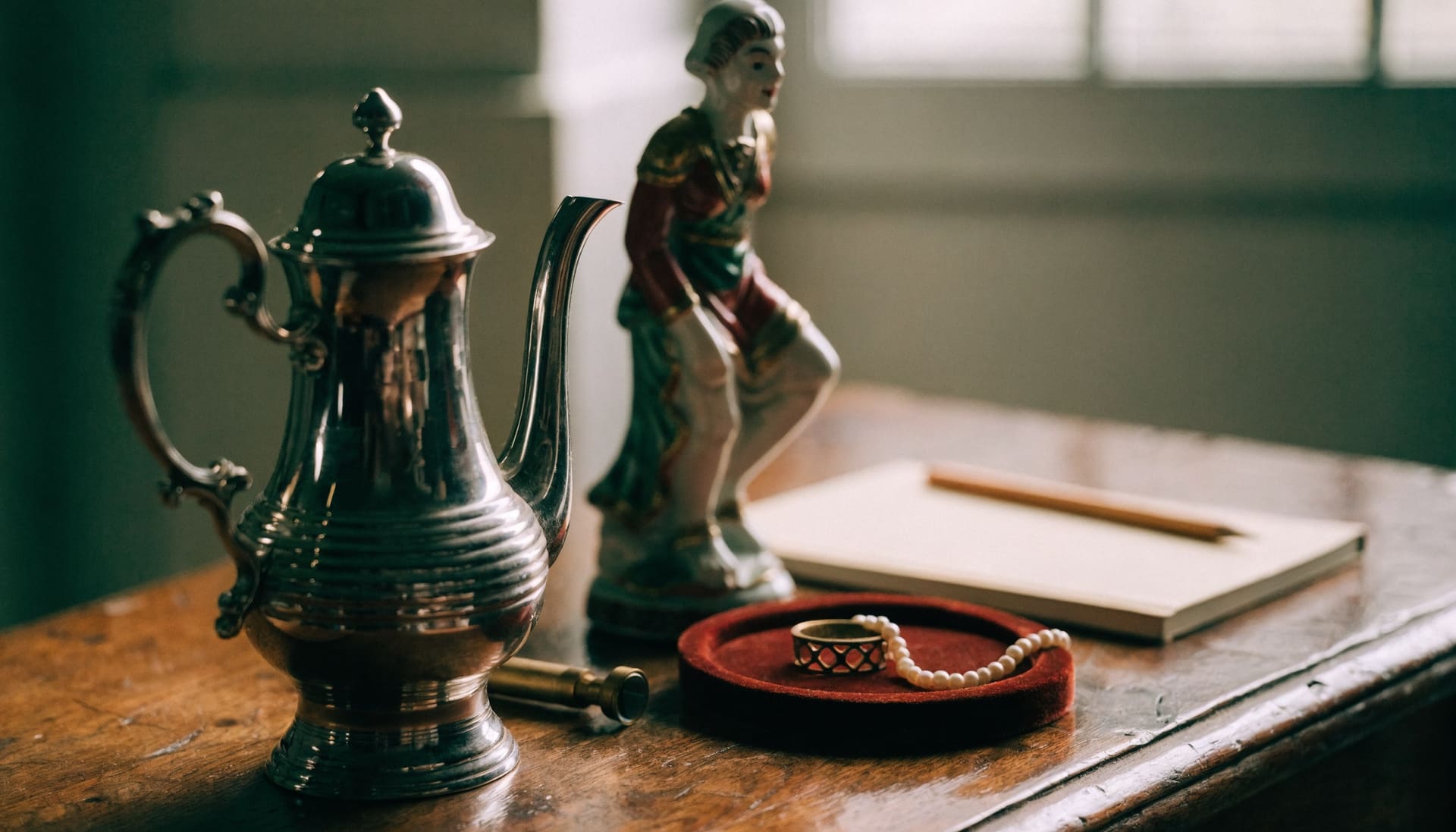 Representative photograph: a silver teapot, porcelain figurine, jewellery and a loupe on an oak desk